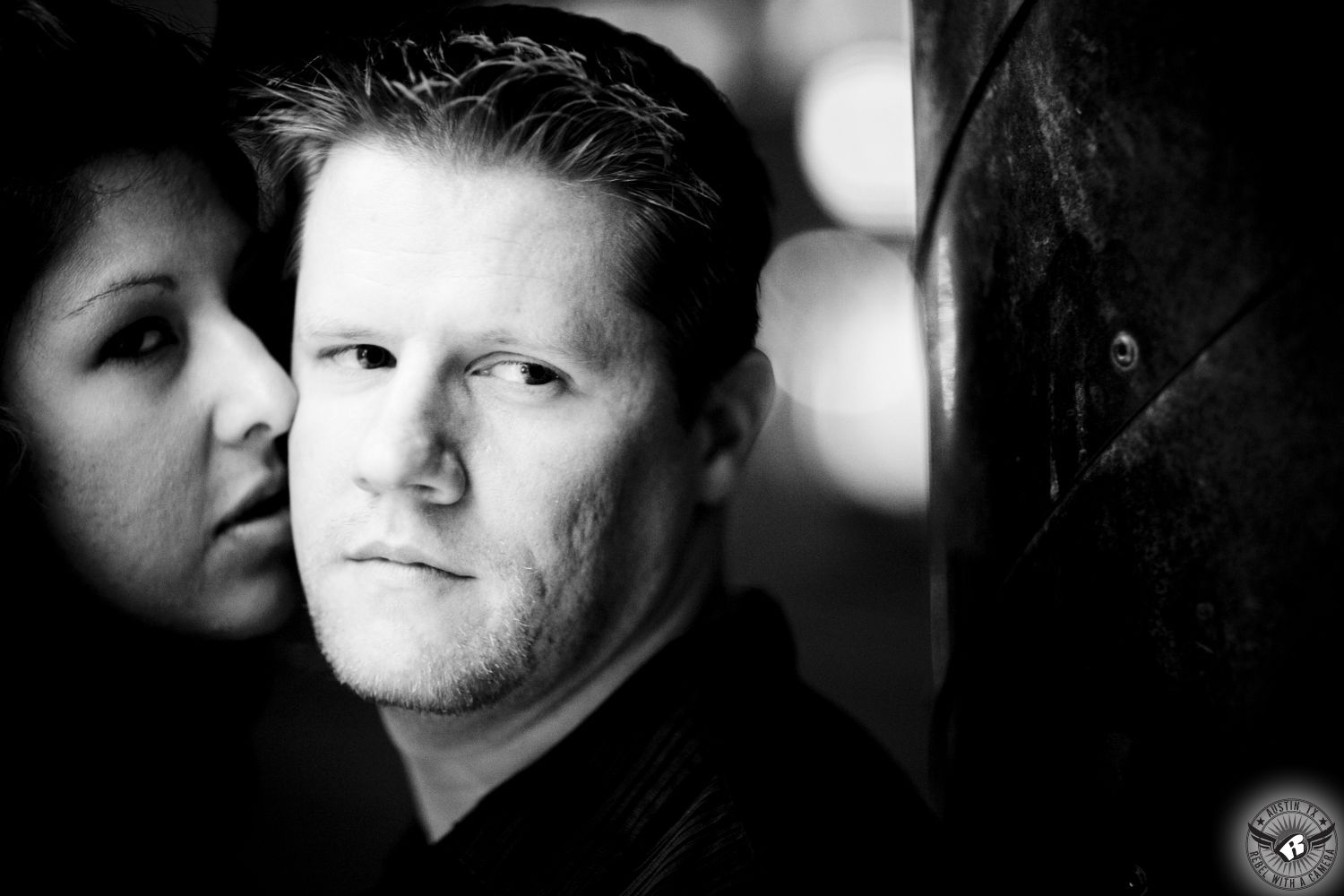 Latino dark haired girl gazes at and nuzzles dishwater blond guy who looks off into distance while leaning on a pole at night with a large boka in the background in this fanciful black and white engagement image on a downtown street in Austin.  